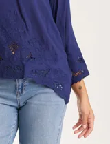 Blusa azul de escote en V y manga larga con terminación amplia. Presenta detalles de bordado y calados tanto en las mangas como en el ruedo delantero.