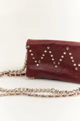 Bandolera color borgoña de cuero con diseño en zigzag de tachas plateadas. Tiene doble asa, una de cuero y otra de cadena metálica plateada.