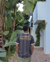 Camiseta de fútbol estilo retro de manga corta, color negro con diseño texturizado a rayas verticales, cuello en V blanco con detalle en mostaza y bordes de mangas a juego. Presenta estampados gráficos en el frente con la inscripción "PLEASURE" y emblemas deportivos.