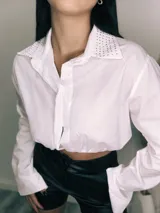 Camisa cropped negra con cuello clásico, mangas largas con puños abotonados y cierre frontal con botones. Presenta detalle de tachas plateadas en el cuello.