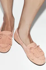 Mocasín plano de piel color rosado, con detalle de flecos y lazo en la parte delantera, costuras visibles y suela plana.
