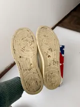 Championes Autry de diseño retro, color blanco con detalles en gamuza beige y suela de tono tostado, con cordones claros y logo lateral de la marca con la bandera de Estados Unidos.