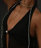 Collar tipo choker y tiras colgantes, compuesto por pequeñas cuentas plateadas. El diseño incluye un choker ajustado al cuello y dos tiras largas que caen sobre el pecho y los hombros, sujetas a una prenda negra con un broche metálico central.