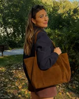 Cartera tipo tote bag de gamuza color marrón, amplia, con doble asa larga para llevar al hombro y bolsillo exterior frontal.