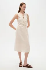 Vestido midi color beige claro, sin mangas y con cuello a la base. Presenta detalles de frunces en el frente, una pequeña abertura en el escote adornada con botones esféricos y corte al cuerpo.