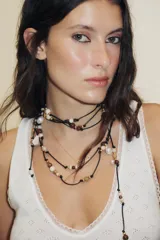 Collar largo de cordón negro ajustable, adornado con múltiples cuentas de piedra en tonos marrones, beige y blanco, y varios dijes metálicos dorados y plateados, incluyendo una concha marina y formas orgánicas.