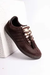 Zapatillas urbanas color tostado con textura geométrica en el cuerpo y detalles de gamuza en la puntera y laterales. Presentan un diseño distintivo con cordones anchos tejidos en tonos tostado y blanco, y un accesorio metálico con borla de gamuza en el empeine.