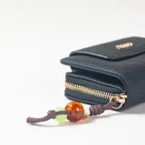 Billetera negra de textura granulada con cierre de cremallera dorada y solapa frontal con broche magnético. Incluye aplique metálico con el logo de la marca en letras doradas y un cordón decorativo colgante con cuentas de colores.