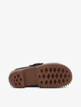 Zapato tipo mule con plataforma track de color marrón y capellada de cuero negro. Cuenta con forro de lana de oveja (borreguito) visible en los bordes e interior, y una correa de cuero marrón con hebilla metálica sobre el empeine.