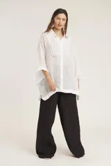 Camisa blanca de manga ancha, con cuello camisero y detalle de pespunte decorativo en forma de 'V' invertida en la parte delantera.