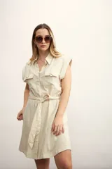 Vestido camisero corto color beige, con cuello clásico, mangas cortas con puños, dos bolsillos con solapa en el pecho, cinturón de lazo y cierre frontal con botones.