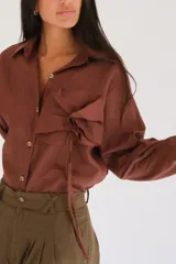 Camisa blanca de manga larga, con botones de guampa y un detalle de roseta o flor a tono en la parte delantera. El calce es holgado con la espalda ligeramente más larga.