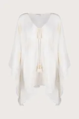 Salida de playa corta tipo poncho en color blanco marfil, confeccionada en algodón ligero y traslúcido. Presenta escote en V con lazo frontal ajustable terminado en borlas y detalles bordados en tono crema.