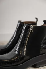 Botas cortas negras con plataforma dentada, confeccionadas en una combinación de cuero con textura de reptil y paneles de gamuza. Presentan un cierre frontal metálico y puntera cuadrada.