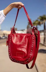 Cartera de cuero color rojo con diseño redondeado, decorada con tachas metálicas plateadas en los laterales y la base. Cuenta con doble asa para llevar al hombro o cruzada, cierre superior y logo grabado en el frente.