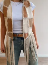 Chaleco de diseño patchwork en tonos neutros con efecto gamuza y pespuntes decorativos, abierto al frente y con largos flecos colgantes en el bajo.