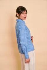 Camisa de manga larga color celeste con rayas verticales blancas, cuello camisero y volados frontales a lo largo de la botonera.