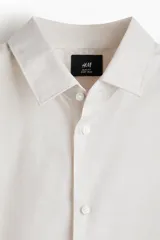 Camisa de vestir color beige claro, de manga larga, con cuello inglés y cierre de botones. Presenta un corte slim fit y bajo ligeramente redondeado.