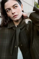 Chaqueta de algodón encerado con diseño de cuadros en tonos verde oliva, beige y líneas rojas. Presenta cuello solapa, cierre frontal con botones a presión metálicos, bolsillos de plastrón con solapa y un detalle de pañuelo desmontable sobre los hombros.