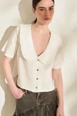 Blusa blanca de lino con escote en V, cuello bobo amplio con detalles de volados y flecos, mangas cortas y cierre frontal con botones de madera.