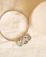 Pulsera rígida plateada tipo esclava, elaborada en alpaca, con tres dijes redondos colgantes. Uno de los dijes tiene grabada la letra 'M'.