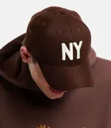 Gorra de seis paneles con ala curva, confeccionada en algodón color marrón. Presenta bordado frontal con las letras NY en color blanco y cierre posterior tipo strapback.