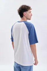 Remera de corte holgado con mangas ranglan en color azul y cuerpo en color blanco. Presenta un estampado frontal con la inscripción 'New York' y detalles gráficos.