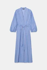 Vestido midi de corte camisero con estampado de rayas verticales en tonos azules y blancos. Presenta cuello con escote en pico, mangas largas abullonadas con puños elásticos y cierre frontal cruzado con botones. Incluye un cinturón del mismo tejido para ajustar a la cintura y bolsillos laterales ocultos.
