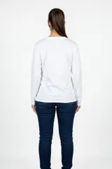 Remera de manga larga color blanco, con cuello redondo y estampado frontal artístico que representa siluetas femeninas en tonos dorados y marrones.