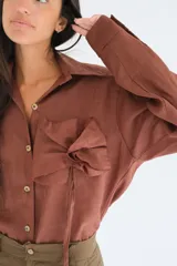 Camisa blanca de manga larga, con botones de guampa y un detalle de roseta o flor a tono en la parte delantera. El calce es holgado con la espalda ligeramente más larga.