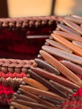 Bolso de mano tejido a mano con tiras de material orgánico en tonos tierra (rojo, marrón, beige) sobre una base de eco-cuero. Posee un asa de mano hecha de un cinturón vintage.