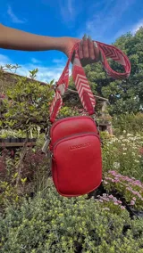 Bandolera compacta color rojo con textura de cuero, correa regulable y desmontable con diseño de rayas rojas y blancas. Cuenta con bolsillos frontales y cierre, interior forrado en tela.