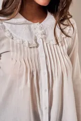 Blusa blanca de algodón peinado con mangas largas, cuello babero bordado y pliegues frontales.
