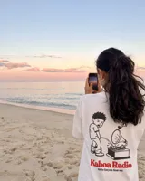 Remera blanca de manga corta con estampado gráfico en la espalda que incluye un personaje ilustrado, un gramófono y la inscripción "Kaboa Radio".
