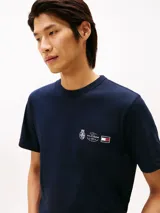 Remera de cuello redondo en color azul marino, confeccionada en algodón regenerativo. Presenta un pequeño logo bordado en el pecho con diseño de escudo y la bandera distintiva de la marca.