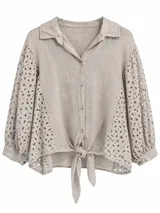 Camisa beige de lino con cuello camisero, mangas abullonadas y detalles de bordado inglés perforado en las mangas y los laterales. Cuenta con cierre frontal de botones y nudo ajustable en la parte inferior.