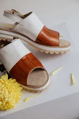Sandalias de cuero con diseño bicolor, combinando una franja de cuero blanco texturizado y una franja de cuero marrón con textura de reptil. Poseen suela track dentada, puntera abierta y tira ajustable con hebilla en el talón.