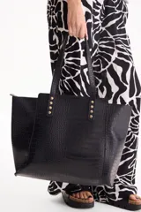 Cartera tipo tote bag de cuero sintético negro con textura croco, doble asa de hombro y detalle de tachas metálicas doradas en la base de las asas.