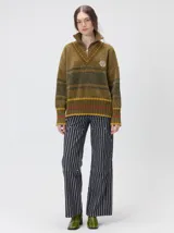 Pullover de textura tipo terciopelo con cuello alto y cierre frontal parcial. Presenta un diseño a rayas horizontales en tonos grises y negros, con detalles en rojo y blanco en el cuello, puños y ruedo. Incluye un aplique bordado con la letra M en el pecho y puños acanalados.