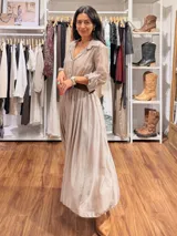 Vestido largo de seda en color beige, con mangas tres cuartos, cuello camisero y falda amplia con caída fluida. Incluye un cinturón tejido en tono marrón que marca la cintura.