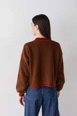 Cardigan de lana color marrón con detalles de bordes en color rojo. Presenta cierre frontal con botones, mangas largas con puños ajustados y un pequeño bordado decorativo en el pecho.