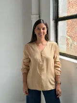 Blusa de color tostado con cuello en V, cierre frontal con botones y mangas largas con puños elásticos. Presenta un diseño de corte recto con bolsillos laterales integrados.