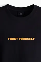 Remera negra de algodón con cuello redondo, mangas cortas y hombros caídos. Presenta un estampado frontal con la frase "TRUST YOURSELF" en color amarillo.