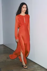 Vestido largo satinado color naranja, con cuello redondo, manga larga con sisa ancha y detalle drapeado con abertura frontal en el pecho. Presenta una abertura en el bajo y cierre lateral con cremallera oculta.