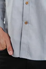 Camisa de vestir blanca de manga larga, con cuello abotonado, bolsillo frontal y botones en tono madera.