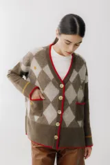 Saco tipo cardigan de punto con diseño de rombos en tonos marrón chocolate y beige. Presenta cierre frontal con botones grandes, cuello en V con borde en color rojo, dos bolsillos delanteros con ribete rojo y detalles bordados pequeños. Posee mangas largas con una franja horizontal en color mostaza cerca del puño.