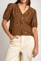 Blusa de viscosa color marrón, con escote en V, mangas cortas abullonadas y cierre frontal con botones. Cuenta con pequeños detalles bordados distribuidos por la prenda.