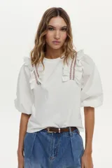 Blusa blanca con cuerpo de jersey y mangas de tela plana abullonadas. Cuenta con volados en los hombros decorados con bordados en contraste.
