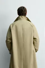 Trench de corte relajado confeccionado en tejido técnico italiano, color beige. Presenta cuello camisero, manga larga, bolsillos de vivo en la cadera y cierre frontal de botonadura simple con botones oscuros.