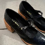 Zapato tipo Mary Jane de cuero vacuno con textura grabada, color marrón oscuro. Presenta una puntera cuadrada, taco bloque de madera y una tira con hebilla metálica sobre el empeine.
